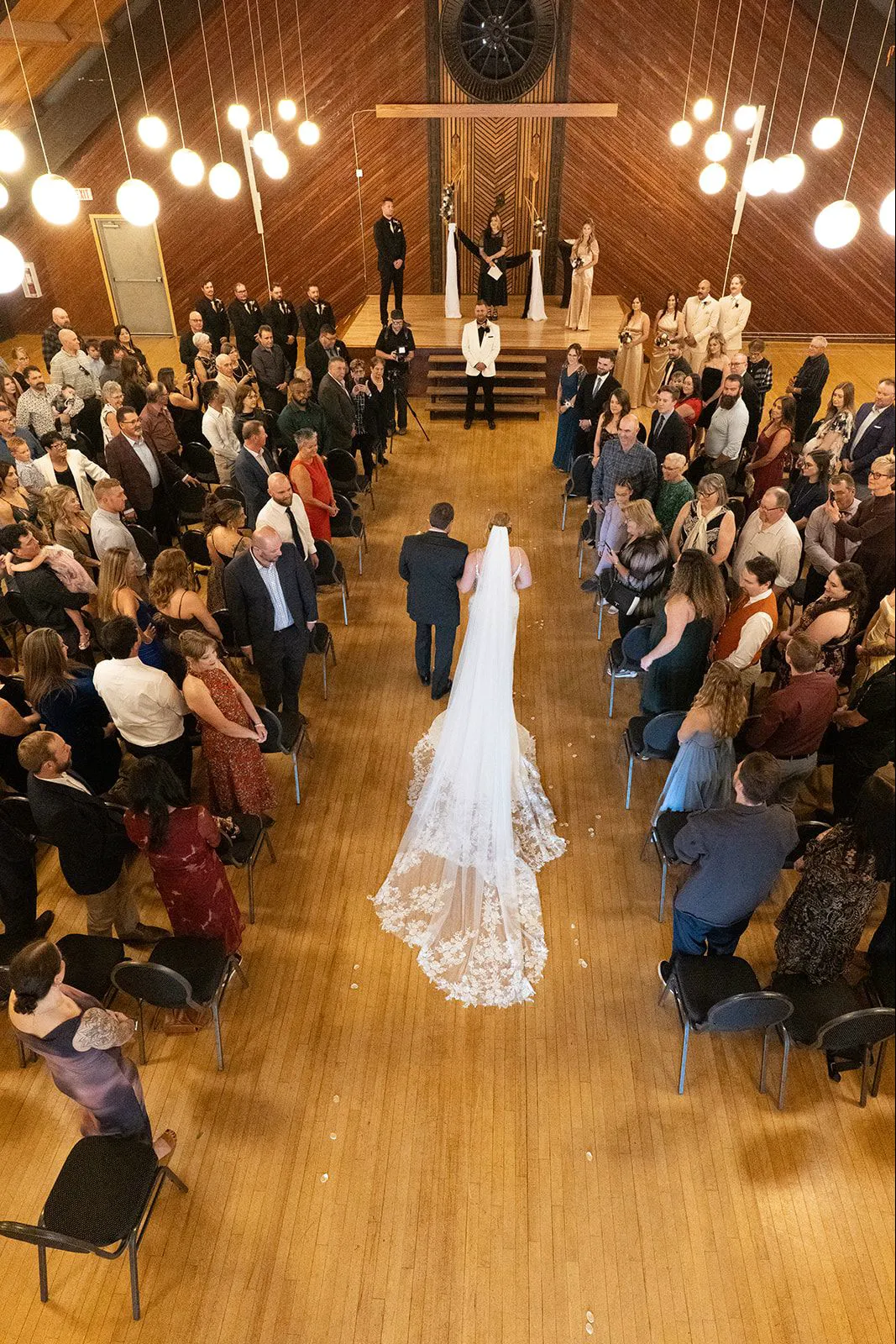 Overhead view of a bride and her father walking down the aisle of a packed community hall